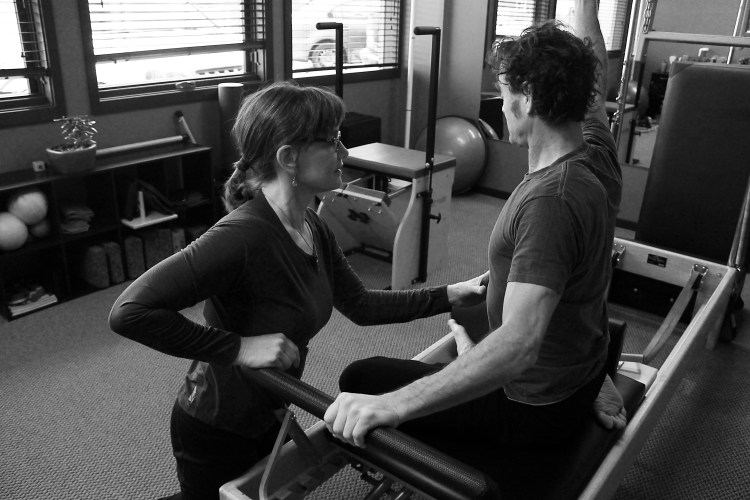 Black and white photo of a man sitting on pilates reformer with arm raised. Woman kneeling in front of him with hand on his ribs